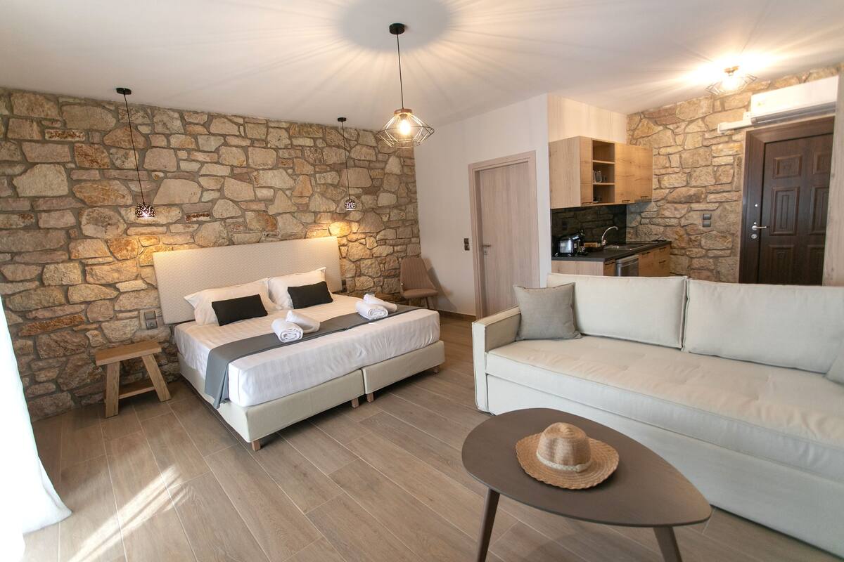 A bedroom with a stone wall, a bed with white linens, a wooden bench, a white sofa, and a small table with a hat.