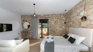 A bedroom with a stone wall, a bed with white bedding, a sofa, a TV, and a dining area with a table and chairs. - Michelangelo Suites and Apartments (Parga)