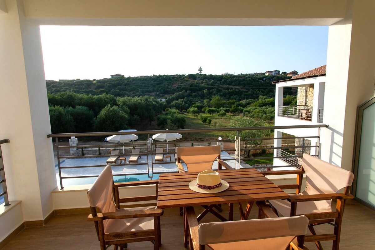 A balcony with a wooden table and chairs, a pool, and a view of a hillside with trees and buildings.