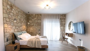A bedroom with a stone wall, a bed with a wooden frame, a bedside table, a TV on the wall, and a large window with curtains. - Michelangelo Suites and Apartments (Parga)