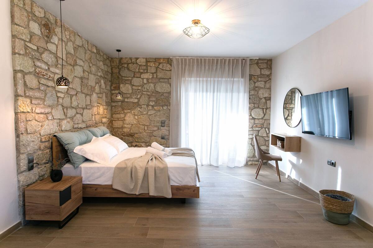 A bedroom with a stone wall, a bed with a wooden frame, a bedside table, a TV on the wall, and a large window with curtains.