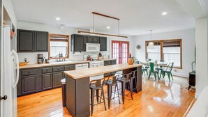 A modern kitchen with dark cabinets, a wooden island with bar stools, a dining area with a table and chairs, and a large window providing natural light. - Secluded Catskill Home w/ Hiking Trails On-site! (Catskill)