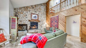 A cozy living room with a stone fireplace, a green sofa, and a wooden staircase. - Expansive Ruidoso Home: Private Hot Tub + Deck (Ruidoso)