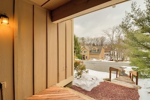 Una terraza de madera con un banco, suelo cubierto de nieve y vistas a una zona residencial con casas y árboles.