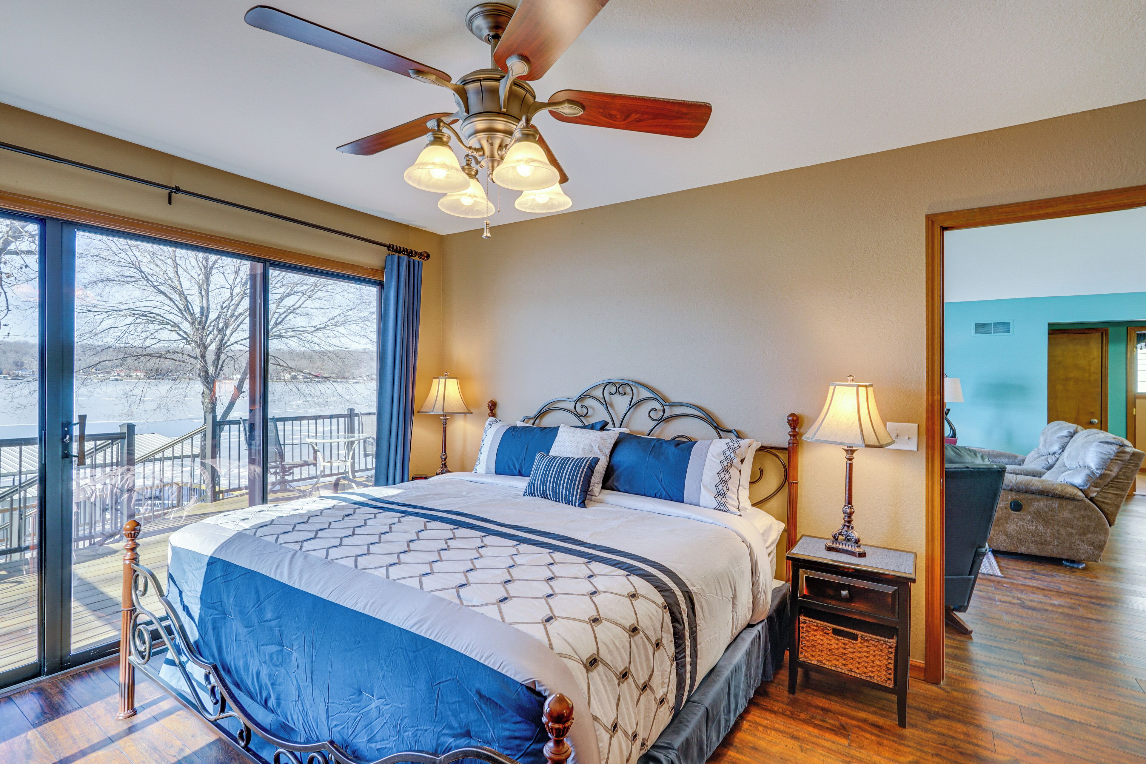 A bedroom with a large bed, a ceiling fan, a nightstand, and a view of a balcony.