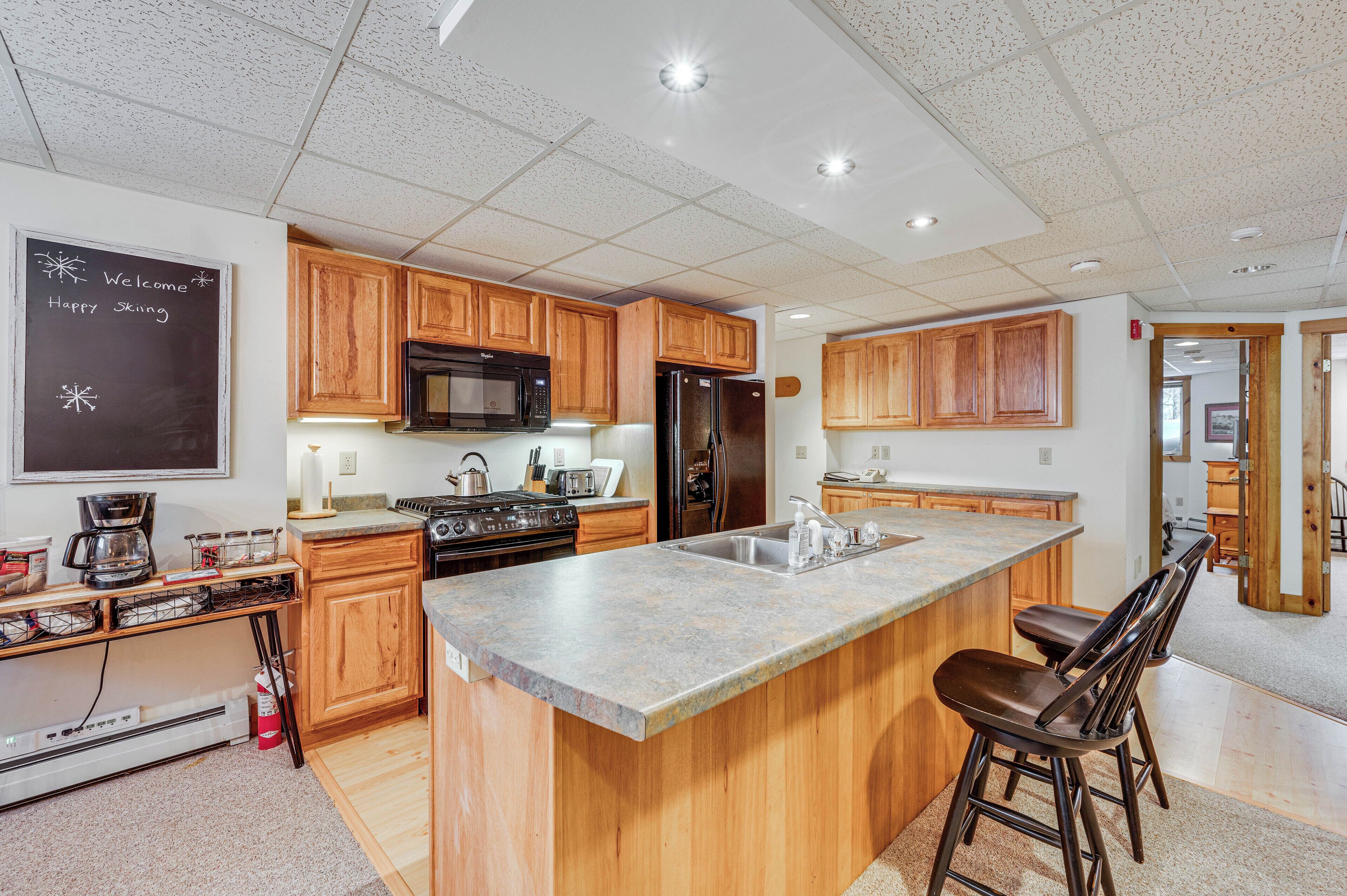 A modern kitchen with wooden cabinets, a large island, stainless steel appliances, and a chalkboard with a welcome message.
