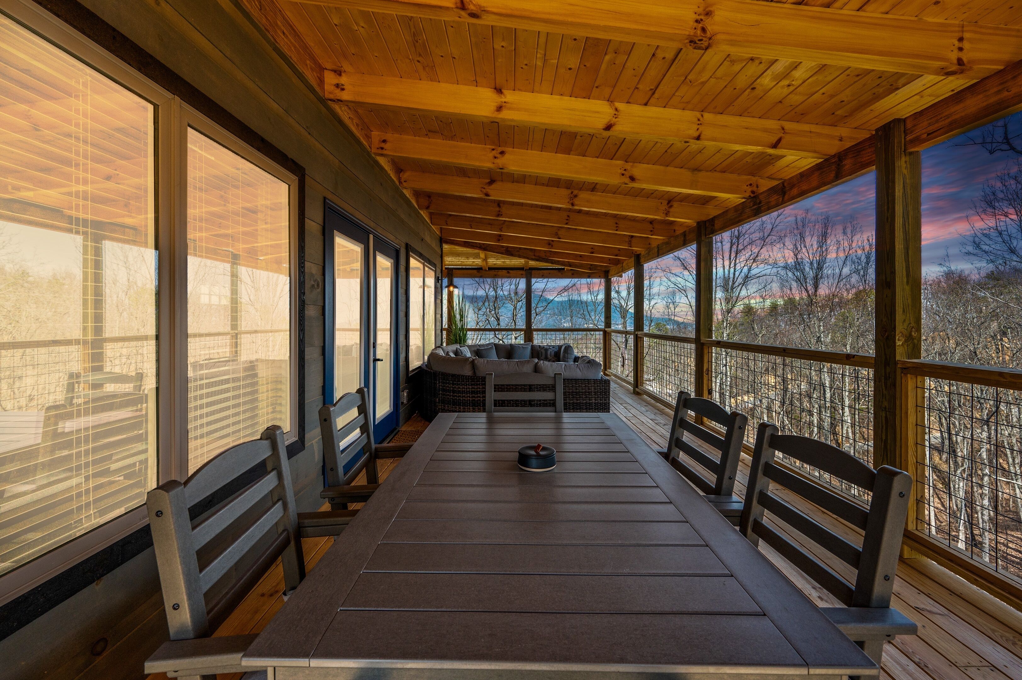 A covered outdoor seating area with a long table and benches, large windows, and a view of trees and sky.