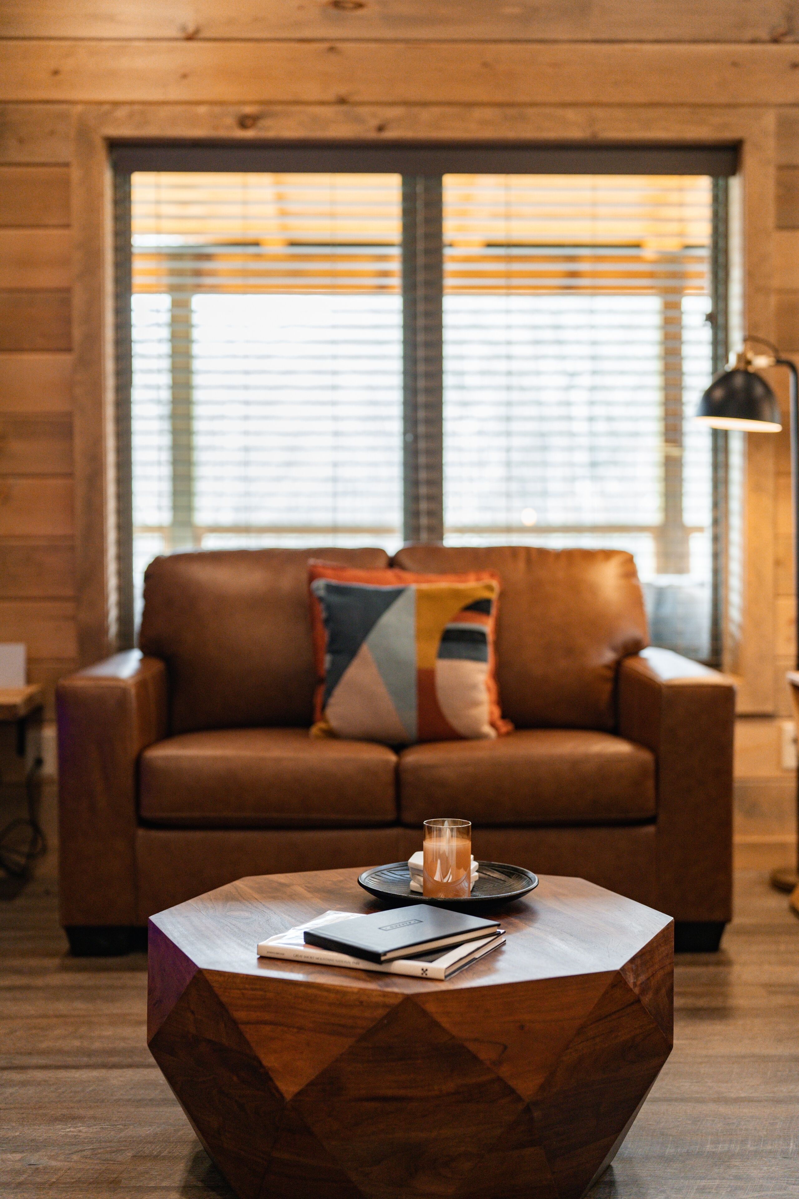 A wooden interior with a brown leather sofa, a geometric coffee table, and a window with blinds.