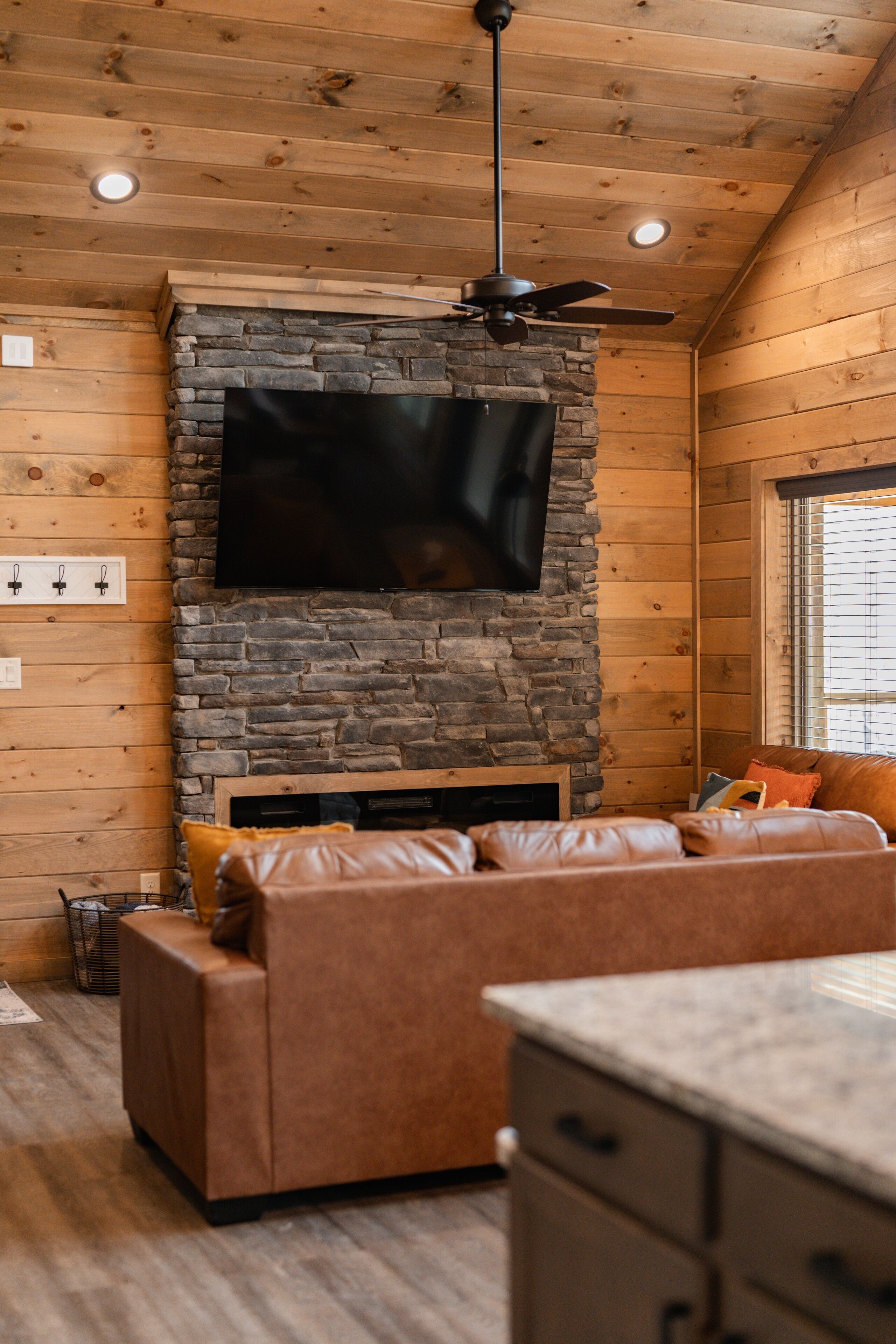 L’intérieur chaleureux d’un chalet avec une cheminée en pierre, un canapé en cuir et un téléviseur à écran plat fixé au mur.