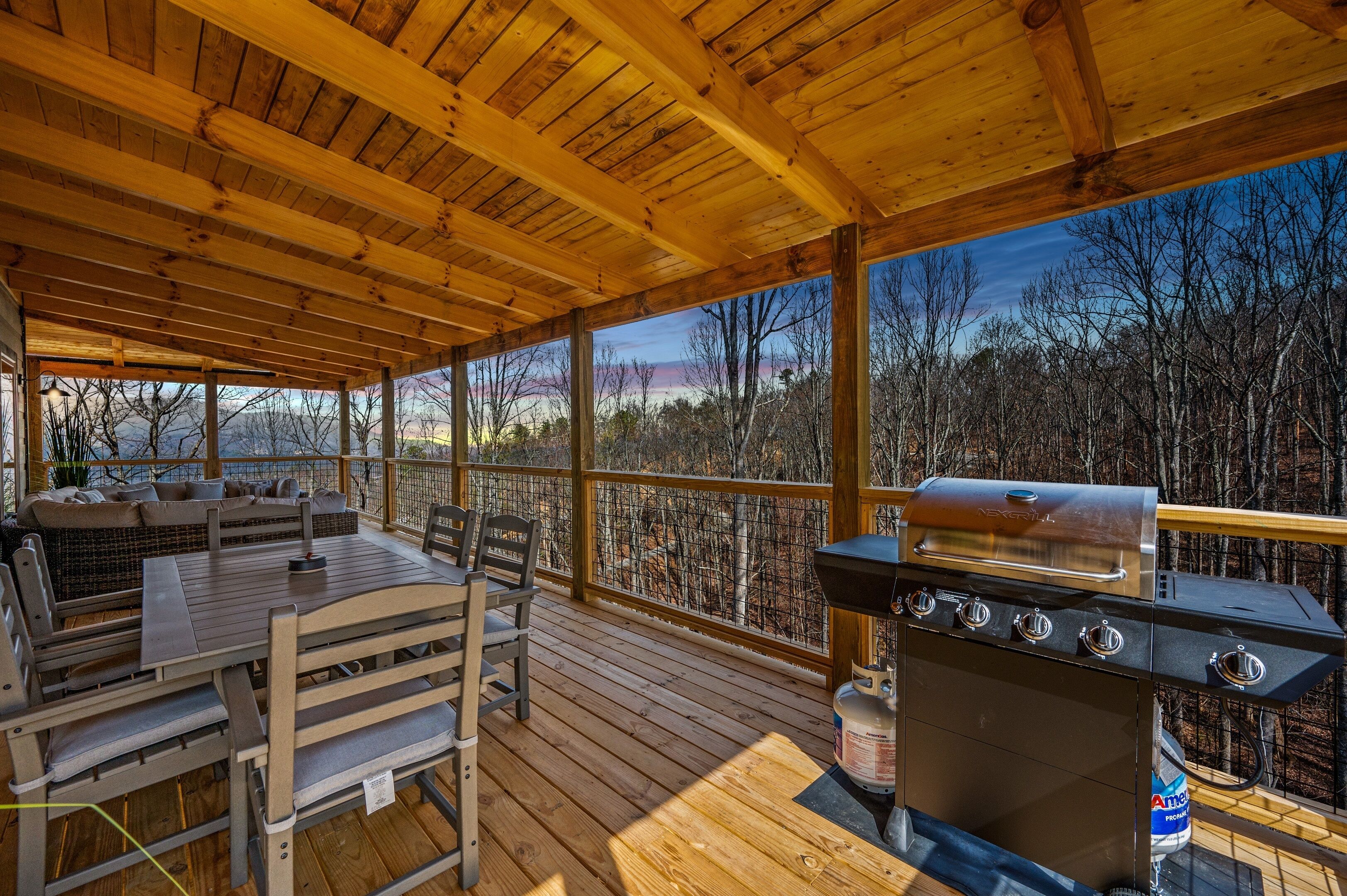 Eine überdachte Terrasse im Freien mit einem Holztisch und -stühlen, einem Grill und einem schönen Ausblick auf Bäume und Hügel.