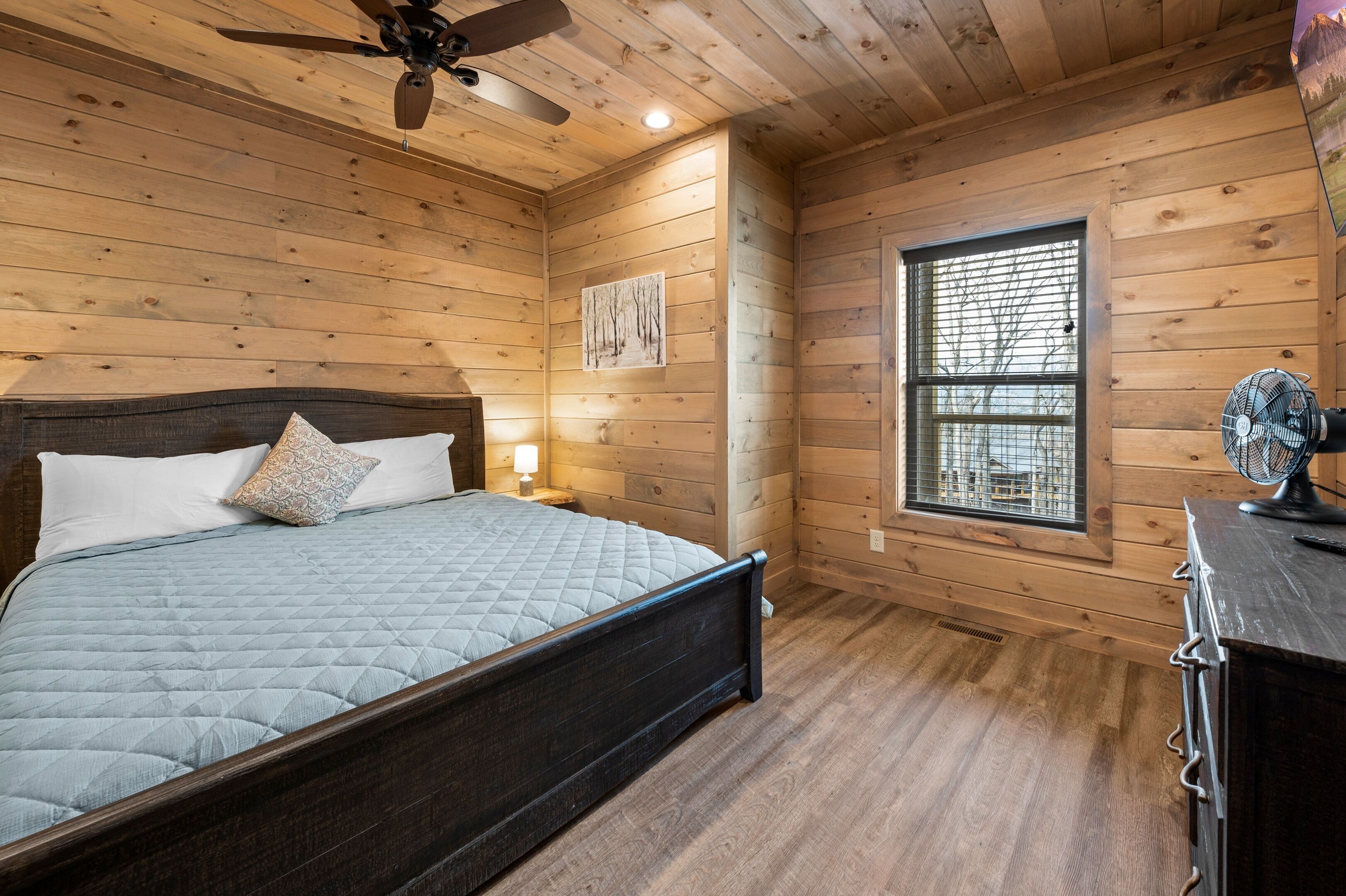 A wooden-paneled bedroom with a bed, a ceiling fan, a window, and a nightstand.