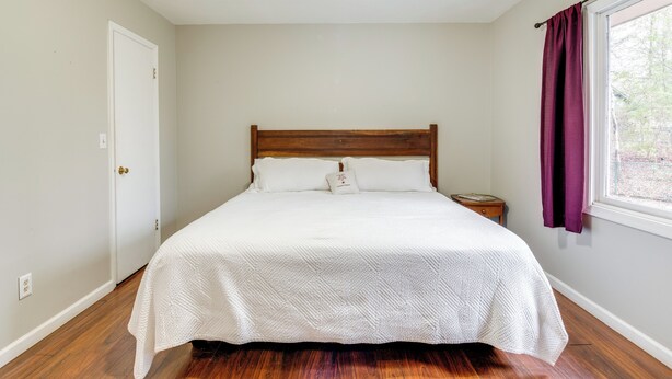 A bedroom with a wooden bed, white bedding, a nightstand, a window with purple curtains, and a ceiling fan. - Pet-friendly Fletcher Home - 13 Mi to Asheville! (Fletcher)