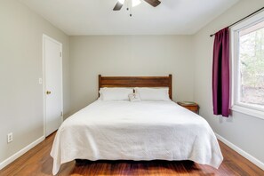 A bedroom with a wooden bed, white bedding, a nightstand, a window with purple curtains, and a ceiling fan. - Pet-friendly Fletcher Home - 13 Mi to Asheville! (Fletcher)