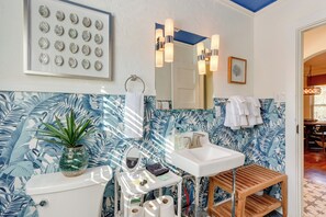 Een badkamer met toilet, wastafel en spiegel. Op het toilet staat een plant, er is een handdoekhouder en een lijst met een afbeelding aan de muur.