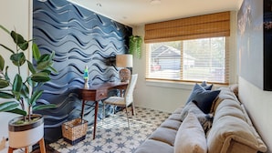 Una sala de estar con una pared azul ondulada, un sofá, un escritorio con una silla, una planta en maceta y una ventana con persianas.