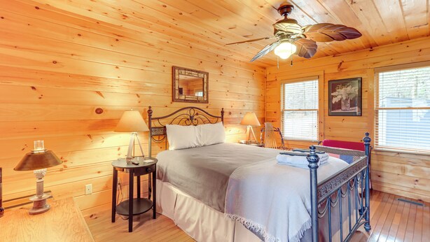 A wooden cabin bedroom with a bed, two bedside lamps, a ceiling fan, and a window with blinds. - Peaceful Cabin w/ Hot Tub: 11 Mi to Blue Ridge! (Epworth)
