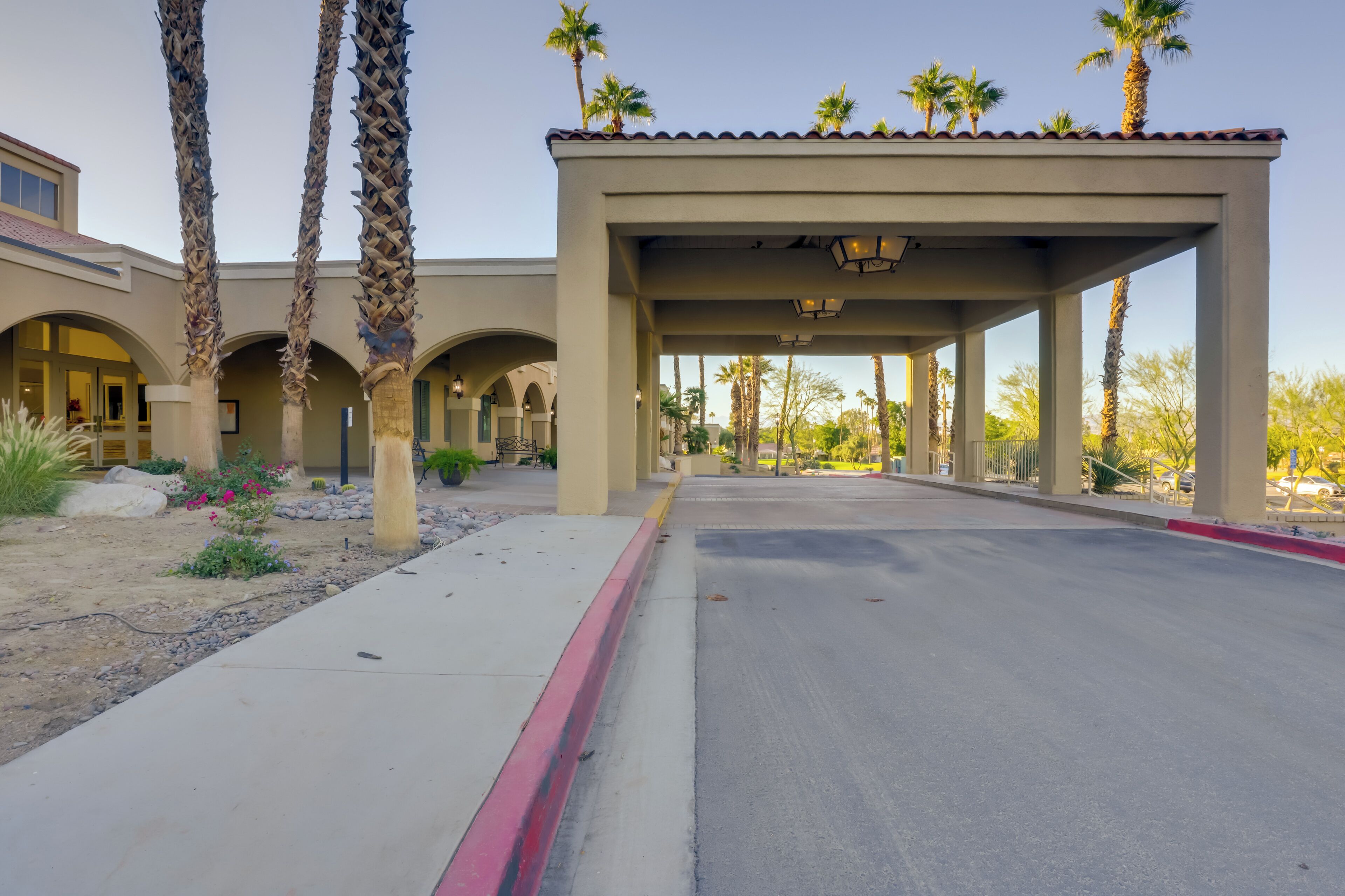 A covered walkway with palm trees, a building with arches, and a clear sky.