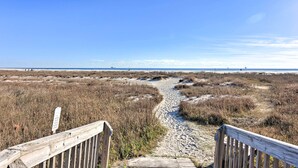 En träpromenad som leder till en strand med torrt gräs och sand.