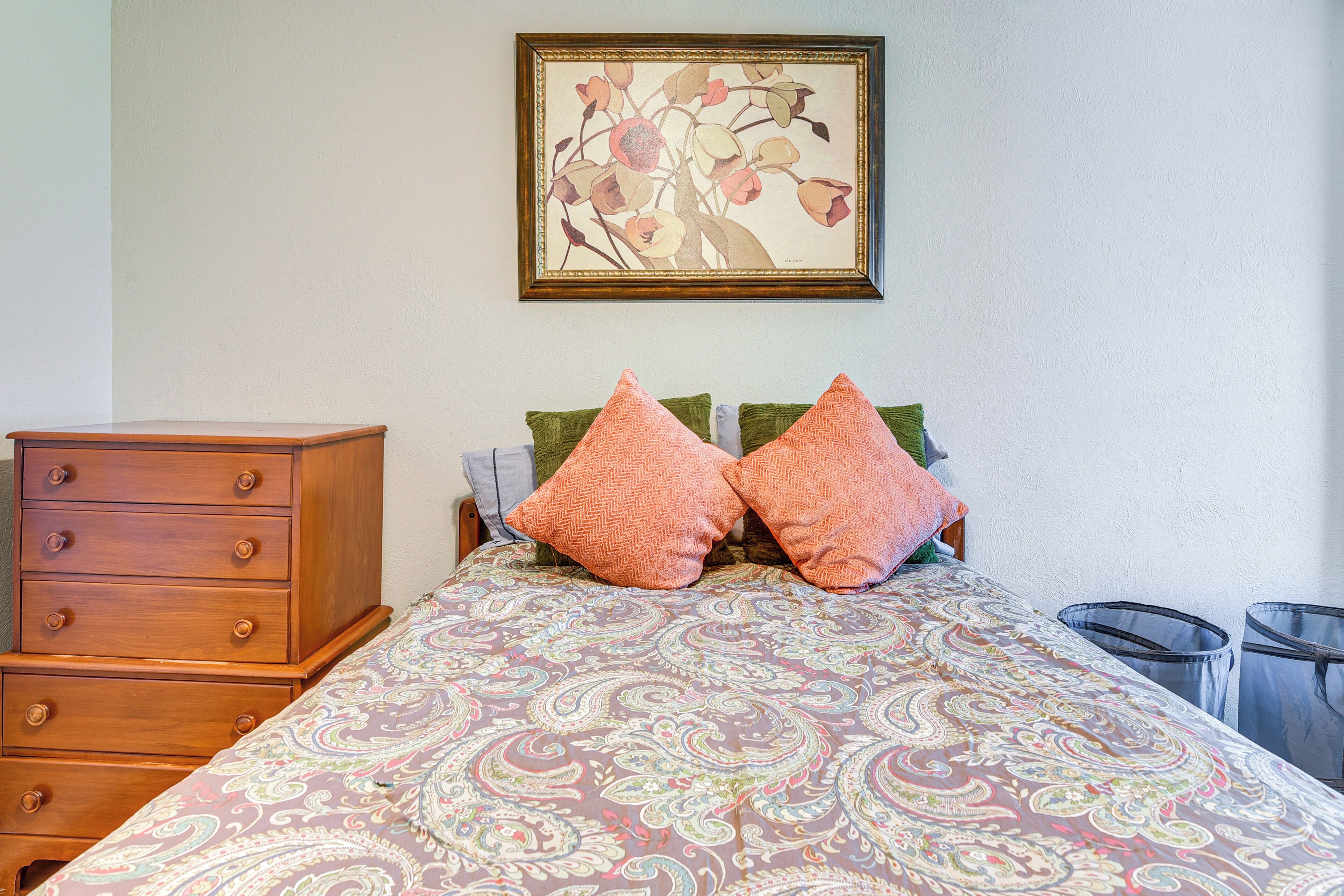 A bedroom with a bed, two pillows, a dresser, a framed artwork on the wall, and two trash bins.