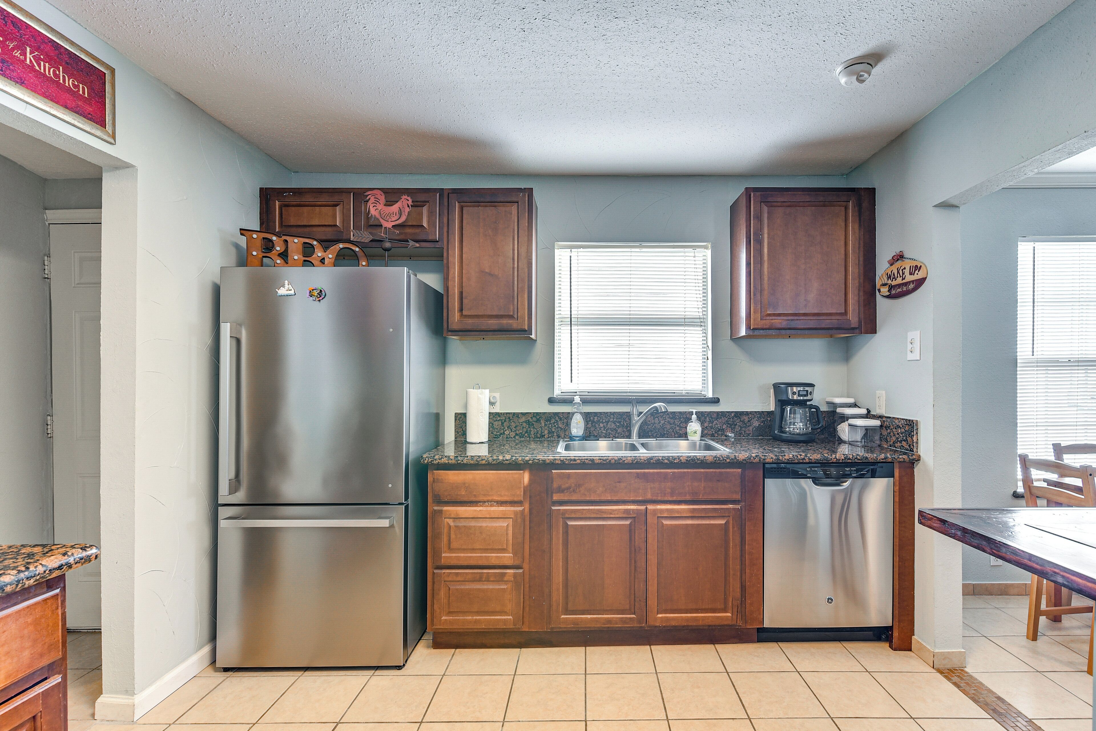 A kitchen with a stainless steel refrigerator, wooden cabinets, a granite countertop, a dishwasher, and a window with blinds.