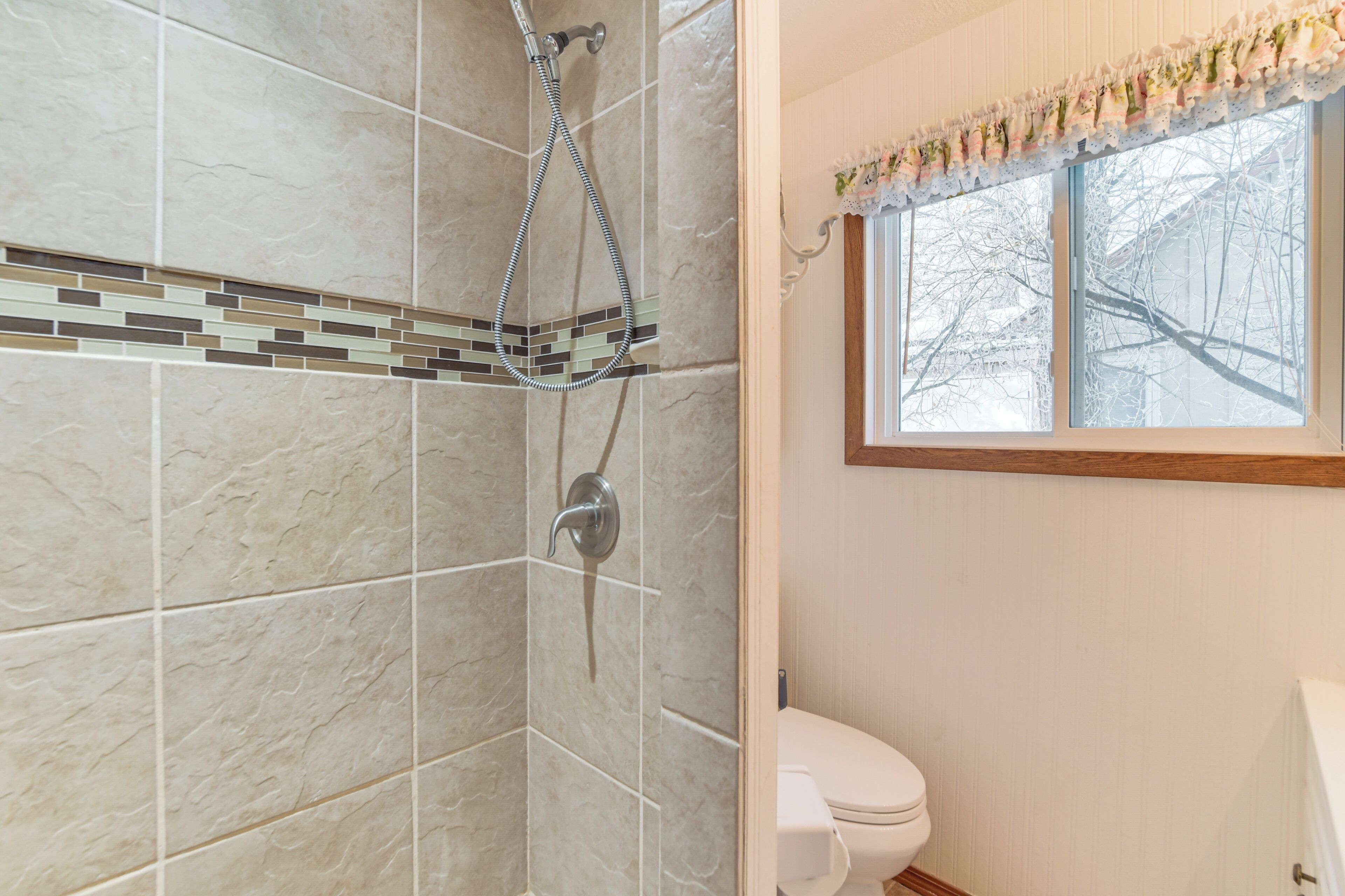 A bathroom with a shower, tiled walls, a toilet, and a window with a view of trees.