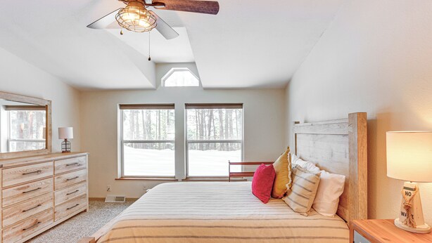 A bedroom with a large bed, a wooden dresser, a ceiling fan, and a window with a view of trees.