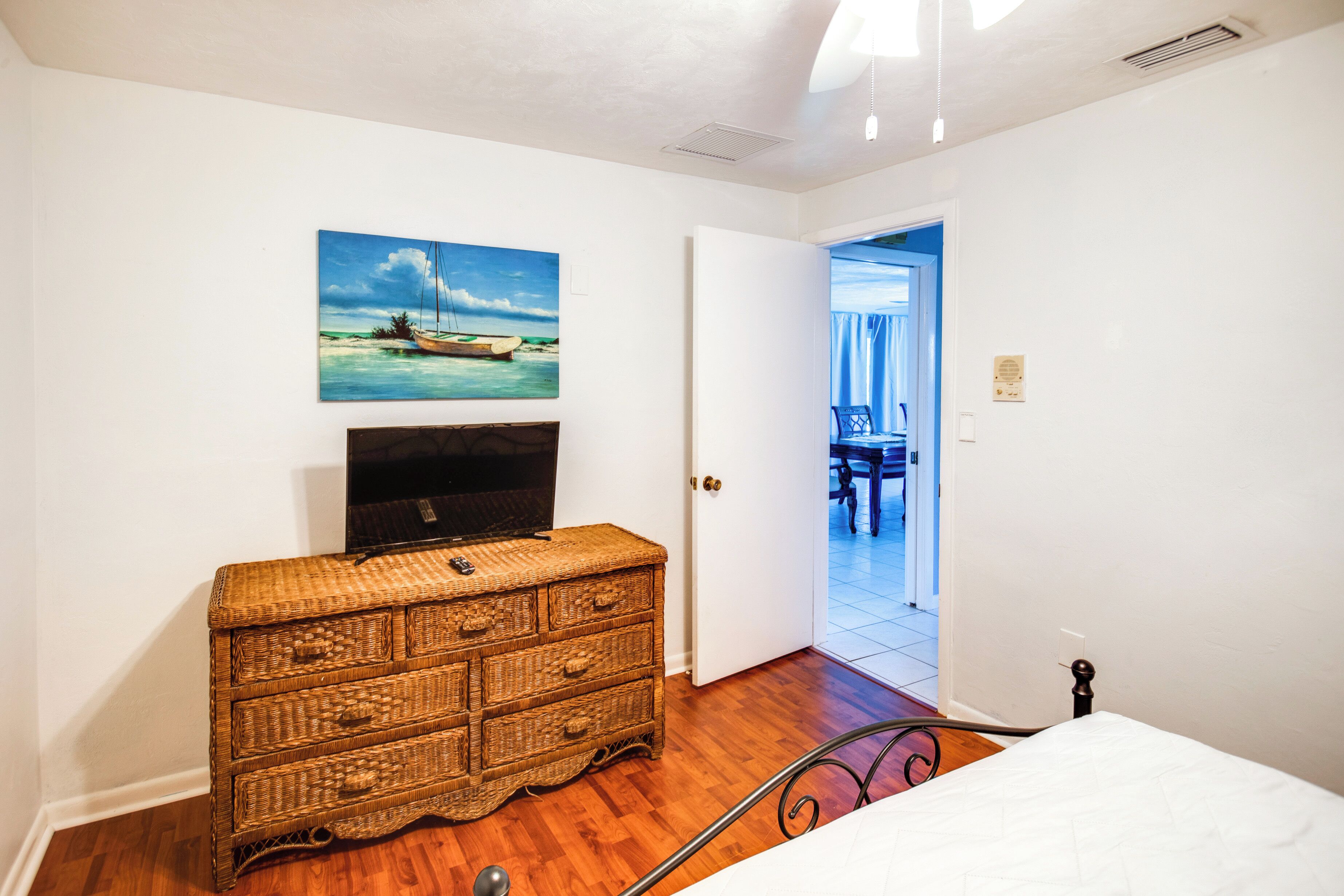 Quarto com cama, cômoda de vime, televisão sobre a cômoda e pintura de uma paisagem praiana.