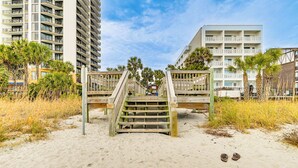 Un escalier en bois mène à une zone de plage au sol sablonneux, où une paire de sandales est posée sur le sable.