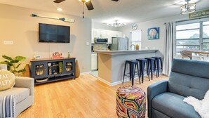 Un salon moderne avec un canapé, un pouf coloré, un ventilateur de plafond et un coin cuisine équipé de tabourets de bar.