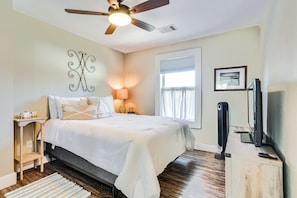 A bedroom with a bed, bedside table, lamp, ceiling fan, window, and a TV stand.
