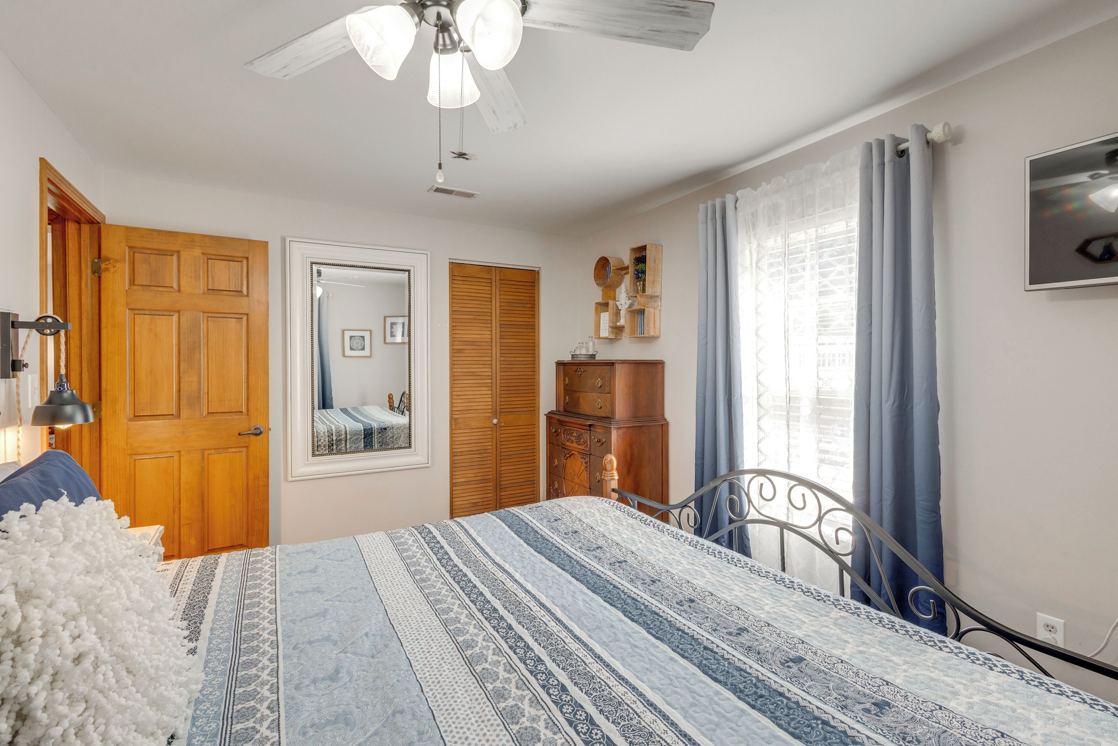 A bedroom with a bed, wooden wardrobe, a mirror, a ceiling fan, and a television on the wall.