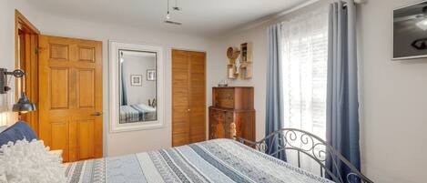 A bedroom with a bed, wooden wardrobe, a mirror, a ceiling fan, and a television on the wall.