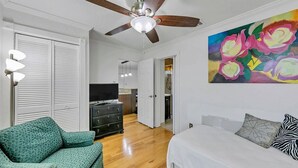 Een slaapkamer met een bed, een groene fauteuil, een plafondventilator, een televisie en een schilderij van bloemen.