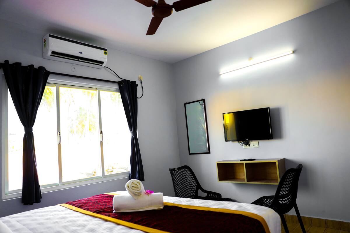Habitación de hotel con cama, televisión, ventilador de techo y ventana con cortinas.