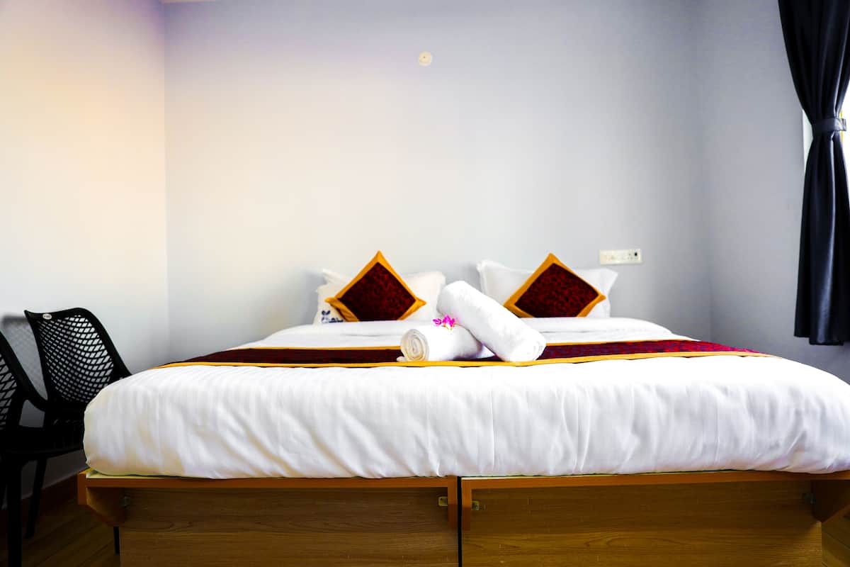 Una cama bien hecha con ropa de cama blanca, dos almohadas con detalles en rojo y amarillo, y una toalla enrollada con una flor rosa.