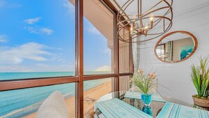 Quarto com uma grande janela com vista para a praia, um espelho redondo na parede e uma mesa com jogos americanos azuis.