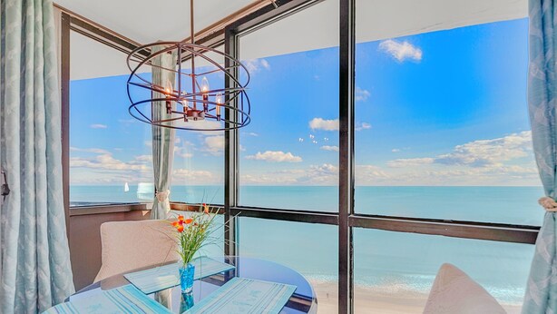 Quarto com uma grande janela com vista para a praia, uma mesa com cardápios e um vaso com flores.