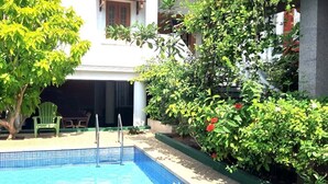 A swimming pool with a tiled edge, surrounded by greenery and a building with a balcony. - Villa Serenity Le Sejour with Swimming Pool (Puducherry)
