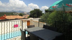 Zona junto a la piscina con una mesa y sillas, piscina cercada y vistas a casas y montañas al fondo.
