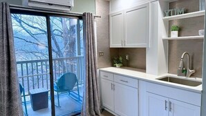 A modern kitchen with white cabinets, a built-in sink, and a window overlooking a balcony with a chair and potted plant. - Apple Valley  56 at Rumbling Bald (Lake Lure)