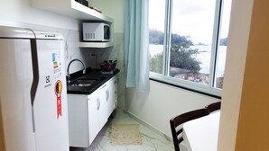 A compact kitchen with a refrigerator, stove, and microwave. A dining table is positioned near a window with a view of a body of water. - Flat Jaguanum (Mangaratiba)