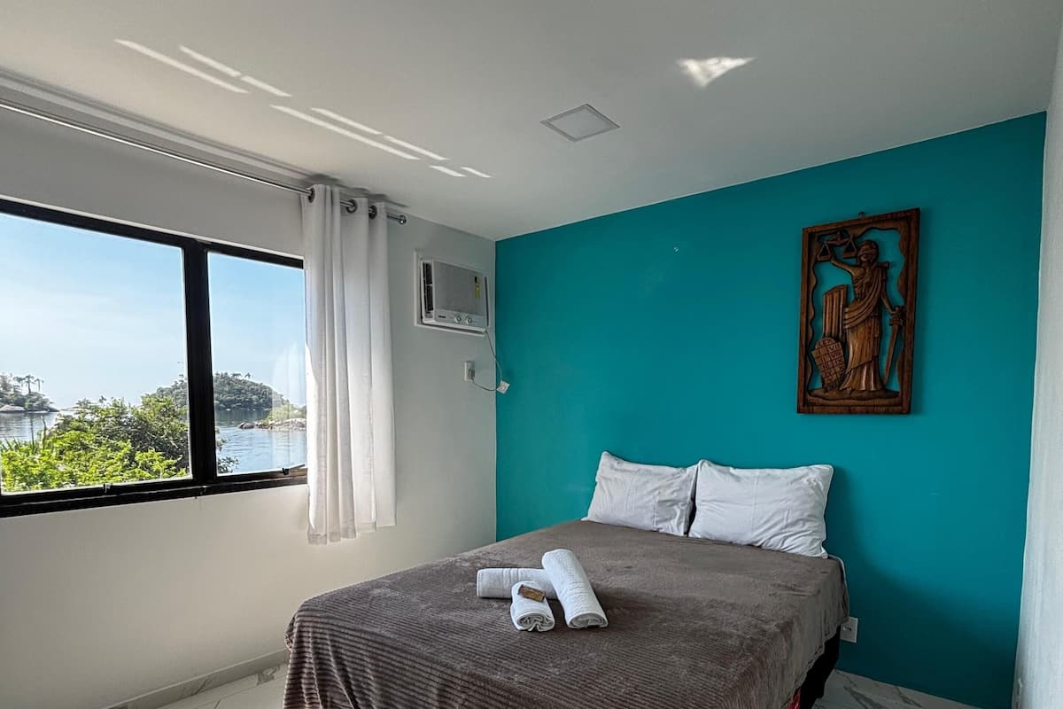Een moderne slaapkamer met een groot bed, uitzicht op zee en een wand in turquoise.