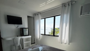 A hotel room with a bed, a desk with a television, a chair, and a window with curtains. - Flat Jaguanum (Mangaratiba)