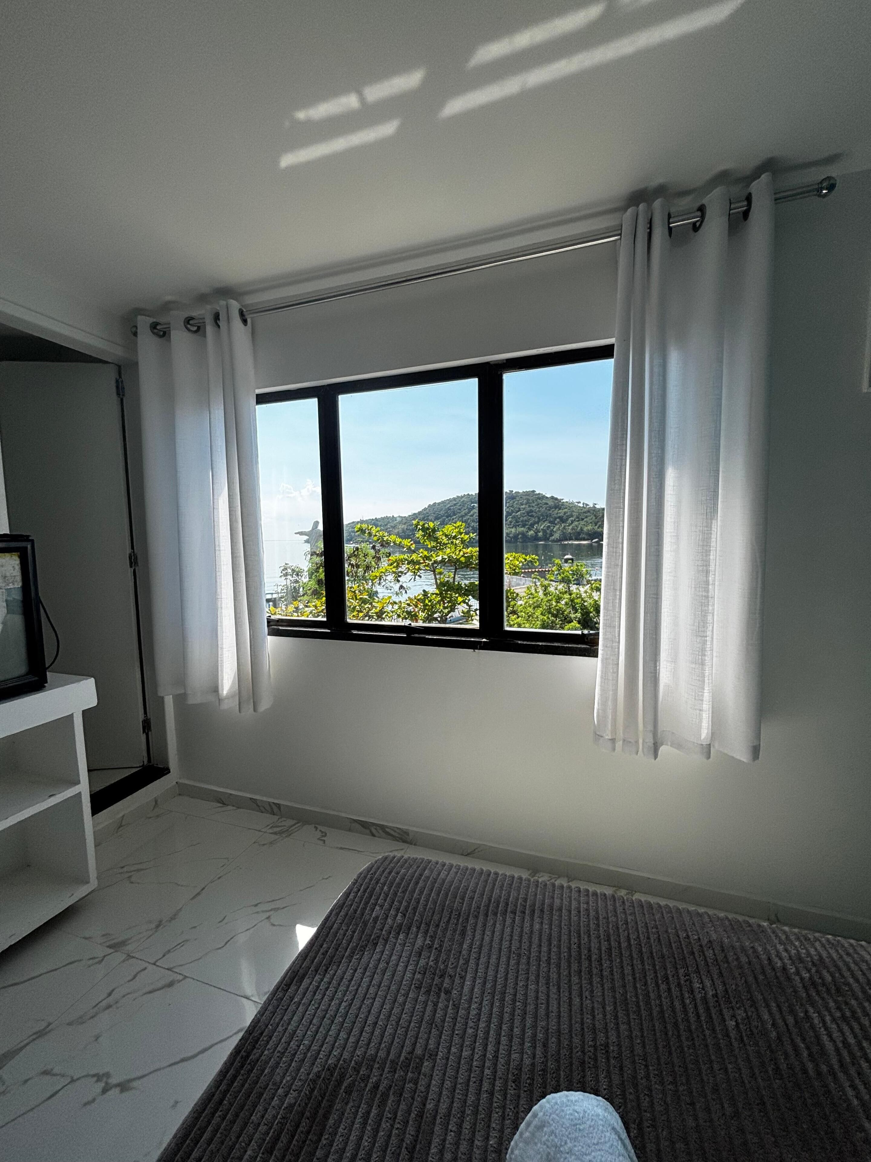 A room with a large window, white curtains, a small table, and a rug.