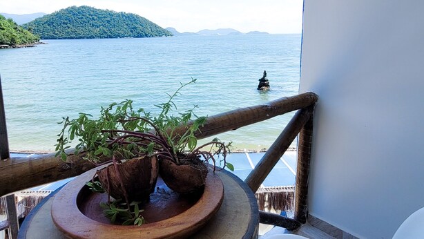 A balcony with a potted plant, a white chair, and a view of a calm body of water with a distant island. - Flat Jaguanum (Mangaratiba)