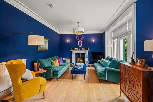 Un salon avec deux canapés de couleur turquoise, un fauteuil jaune et un meuble en bois. Une cheminée surmontée d'un lustre rose se trouve au centre de la pièce.
