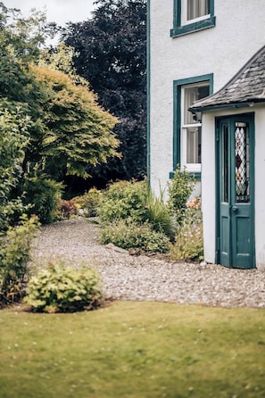 Un bâtiment blanc avec des bordures vertes et une porte, entouré d’un chemin de gravier et de diverses plantes.