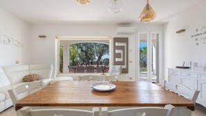 Een moderne eethoek met een houten tafel en witte stoelen, een witte bank en uitzicht op bomen door een glazen deur.