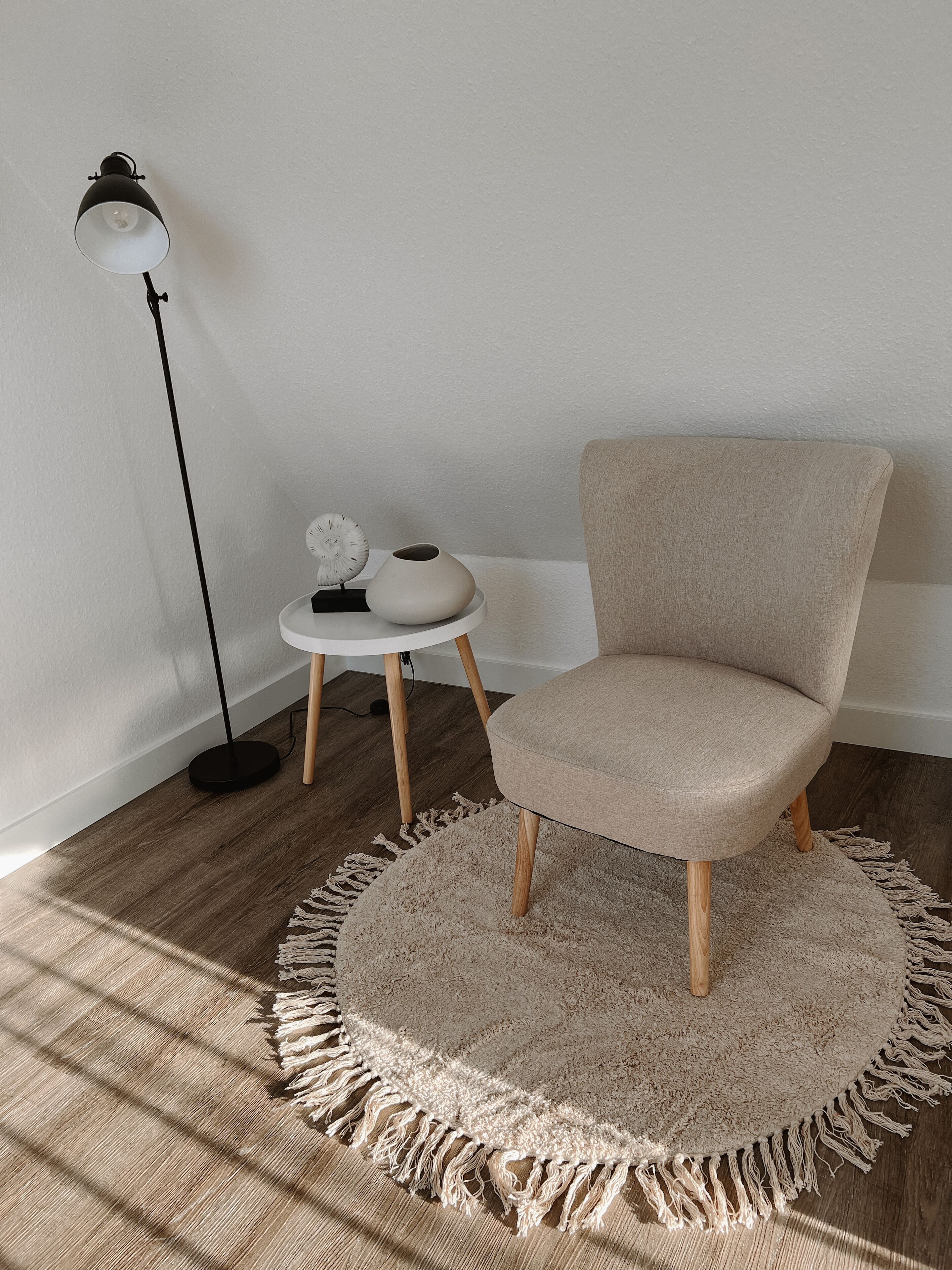 Een kamer met een rond vloerkleed, een stoel, een bijzettafeltje met een vaas en een klok, en een vloerlamp.