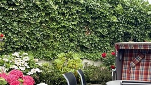 An outdoor patio area with a table and chairs, surrounded by a hedge and flowering plants. - Residenz zum Südstrand - Kutte (Borkum)
